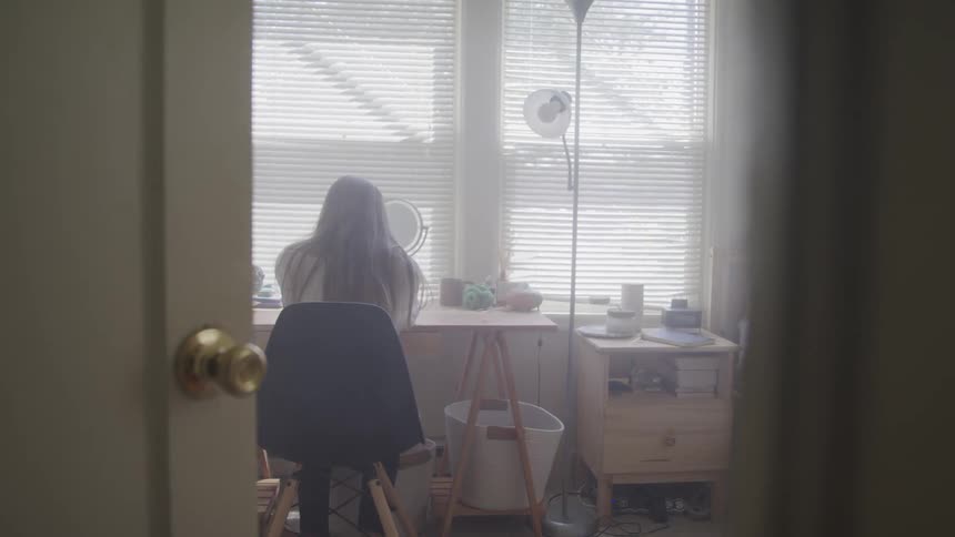 A woman sits at a desk in a bedroom at a table with a makeup mirror on it