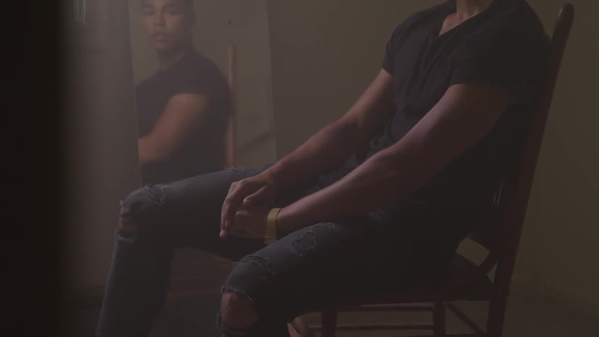 A young black male sits on a chair in a dimly lit room and looks at himself in a mirror