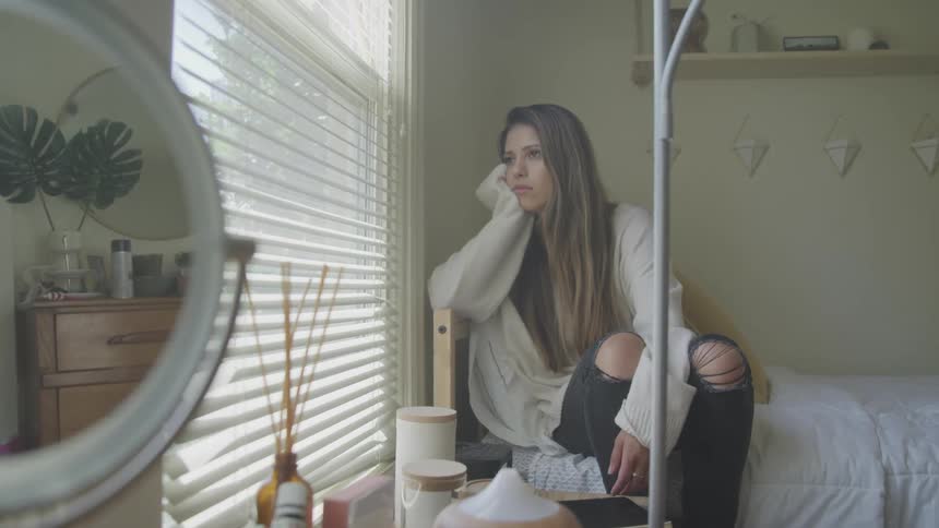 A woman sits on her bed leaning on her hand looking out her window