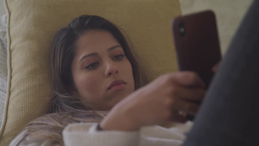 Brunette woman lying on the bed uses smartphone in her room