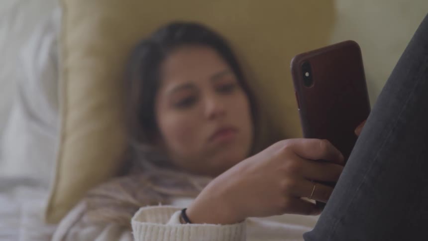 A young girl lies in her bed while looking at her phone