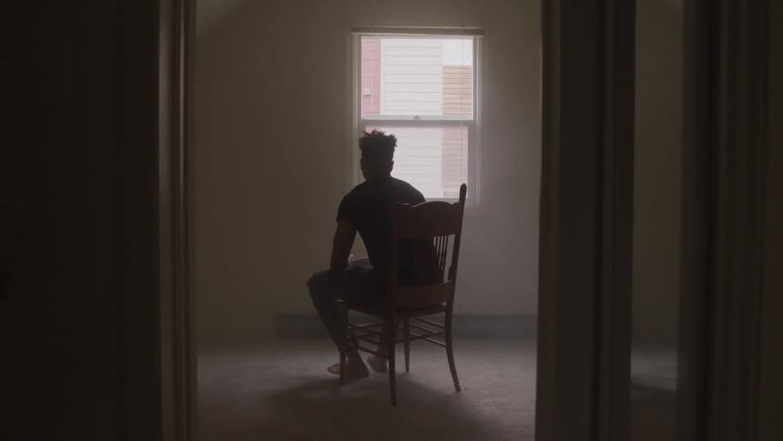 A young man sits in a chair and looks out a window