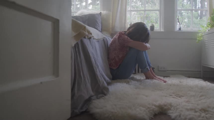 A sad female sits on the floor of a bedroom with her head and arms on her knees
