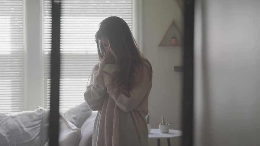 A young girl stands in a room and puts her hands up to her face as if crying in to them