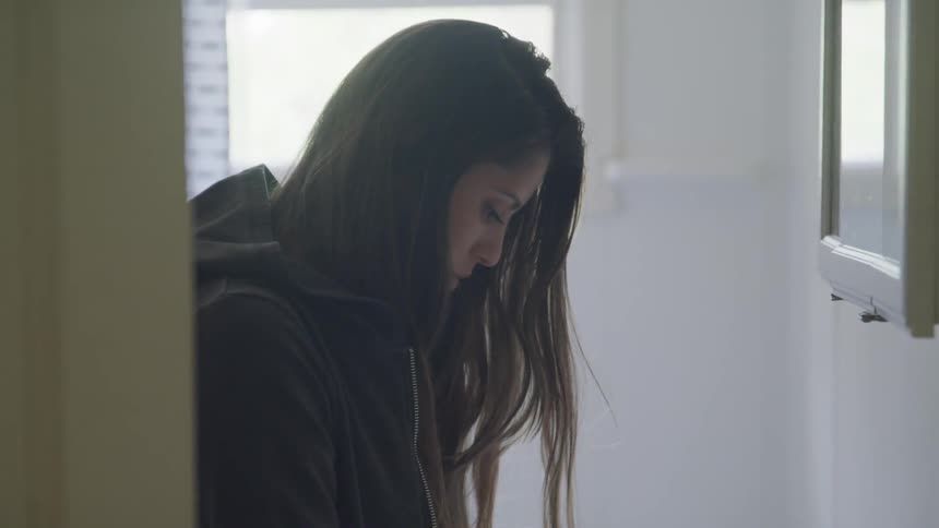 A young woman sits in a small room looking downward without much emotion