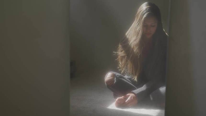 An unhappy young woman sitting on a floor before a mirror in an otherwise empty room