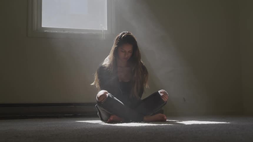 A female teenager sits cross legged on the floor with her eyes down