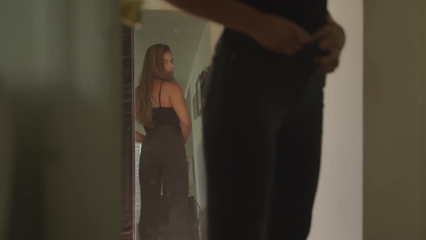 Woman looks at herself in a mirror as she does up her pants