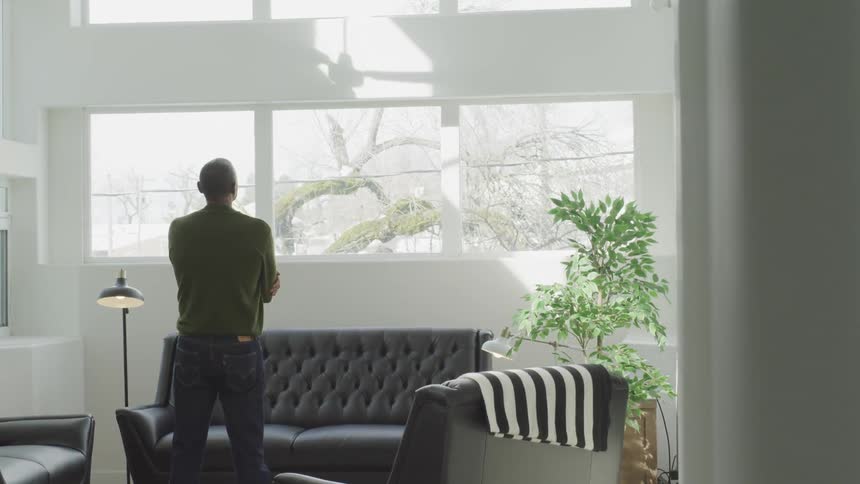A man is standing in a living room with his arms crossed in front of him as he looks out the windows spanning most of a wall behind the sofa