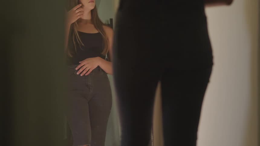 A woman smooths her clothes and strokes her hair as she looks at her reflection in a full length mirror