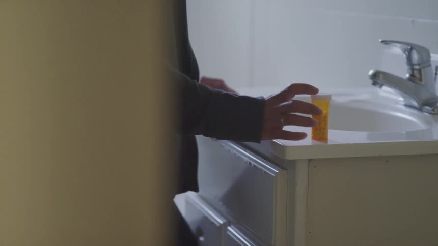 A woman stands at a sink and reaches for a bottle of pills that is on the counter next to it