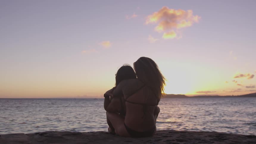 A woman hugging a person from behind on the beach while they look at a sunset