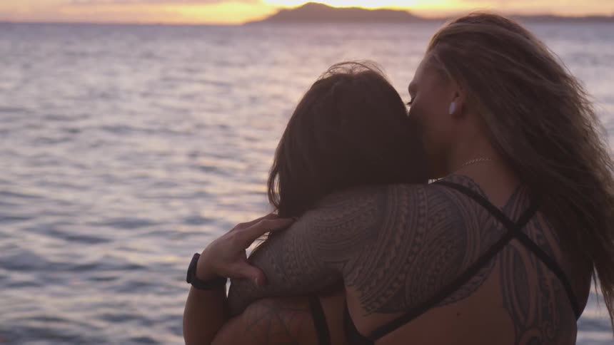 A woman with tattoos with her arms around another as they watch the sunset on the ocean
