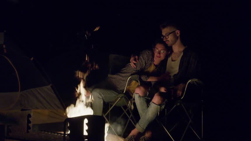 Man and woman cuddle as they sit near a campfire at night