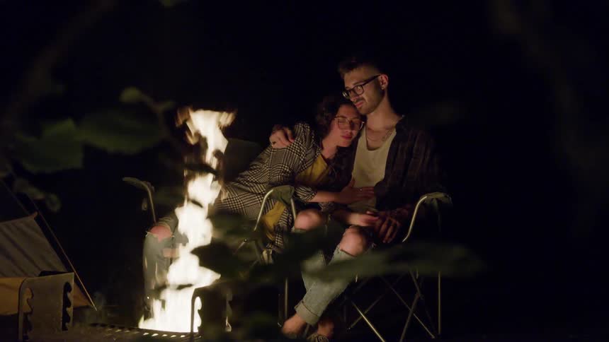 Man and woman sitting on the chairs hold each other behind the campfire in the forest at night