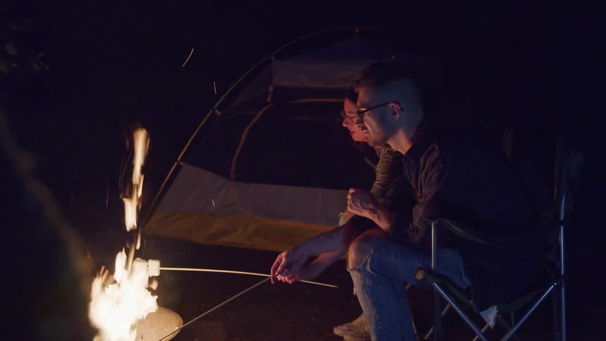 A young man and woman sit outside and roast marshmallows over an open fire