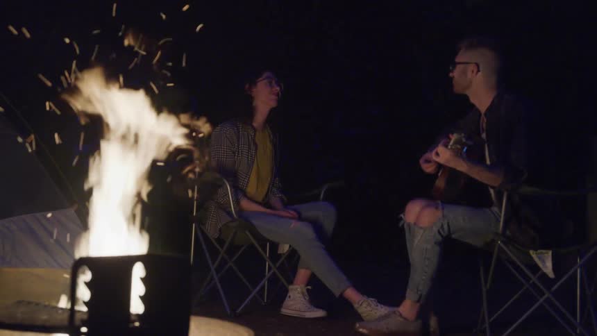 Couple sitting by a fire pit relaxing and playing guitar while camping