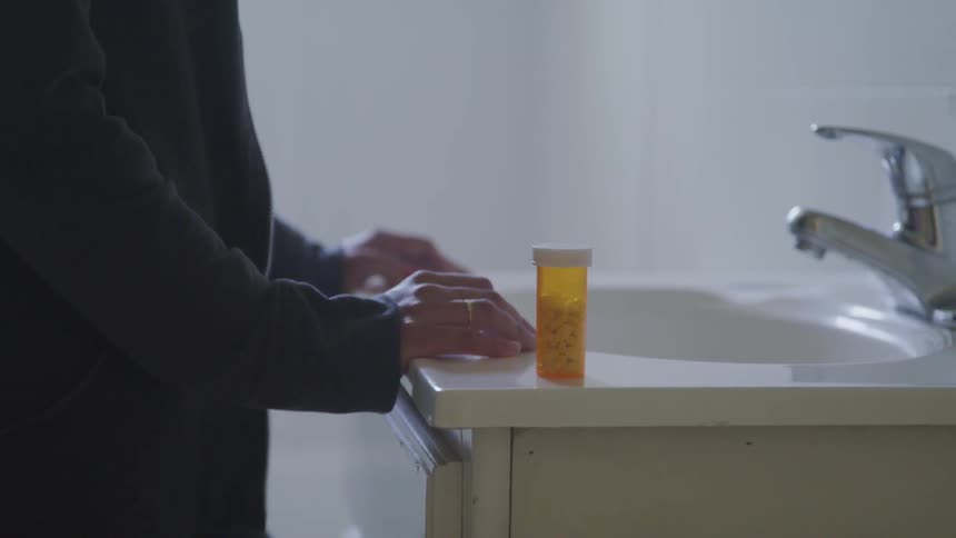 A man stands with his hands on a bathroom counter next to a bottle of pills