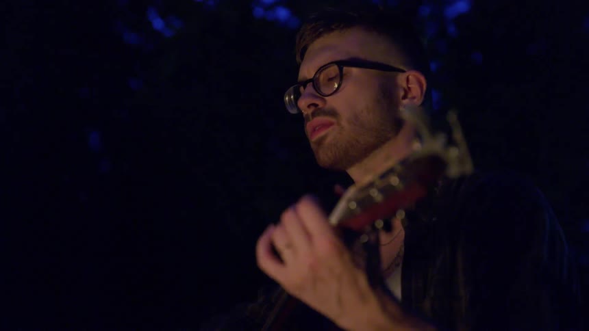 Man wearing glasses sings as he plays a guitar