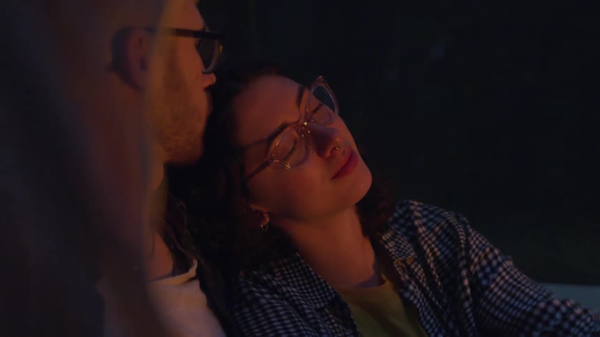 A white female is leaned against a white male as they sit in front of a bonfire outside and the man kisses his significant other on the head
