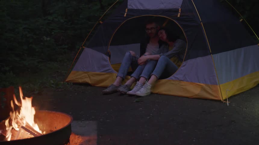 A man and woman sit in a tent while a campfire burns in front of them