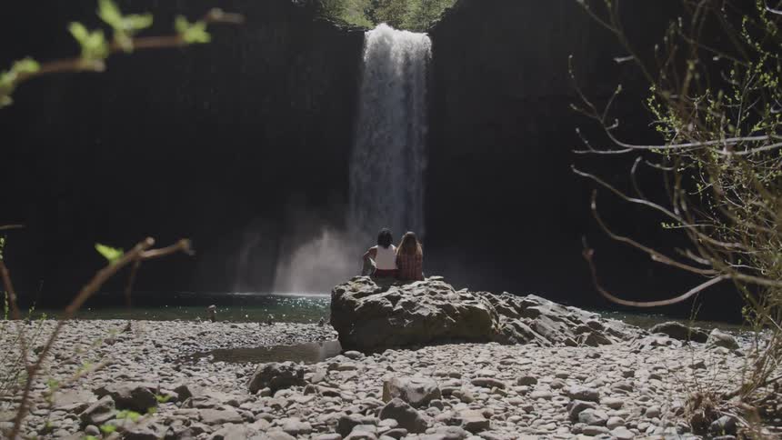 Black man and woman sitting on the big rock looks at the waterfallin the forest