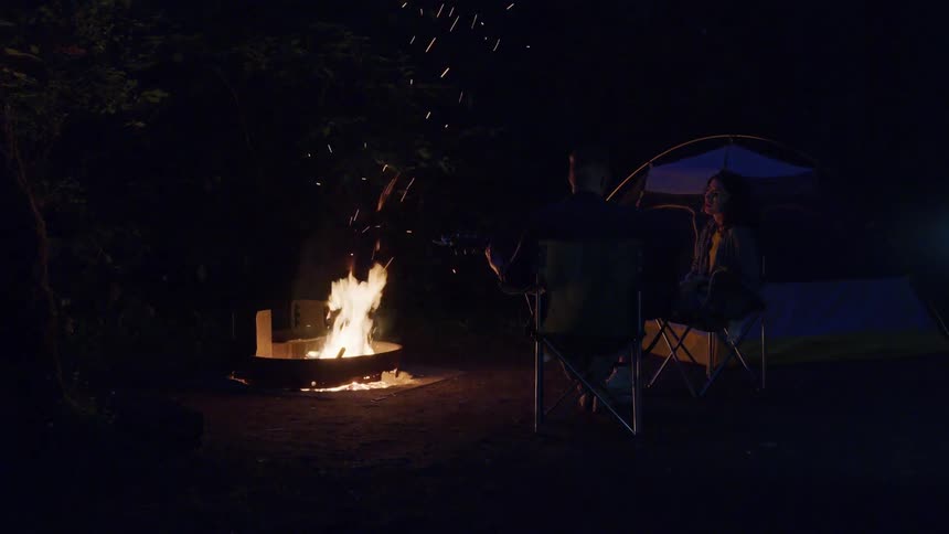 A couple sits on chairs outside their tent as embers fly up from a campfire at night
