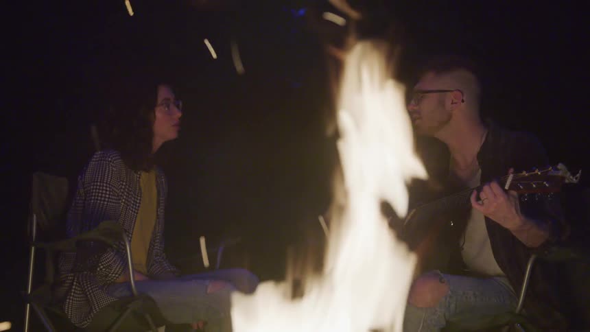 A man and woman sing and play guitar around a fire