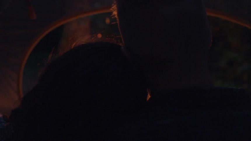 Woman rests head on mans shoulder outdoors near fire