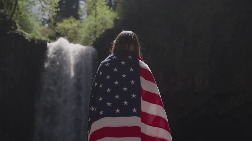 Woman wearing american flag on her shoulders looks at the waterfall in the forest