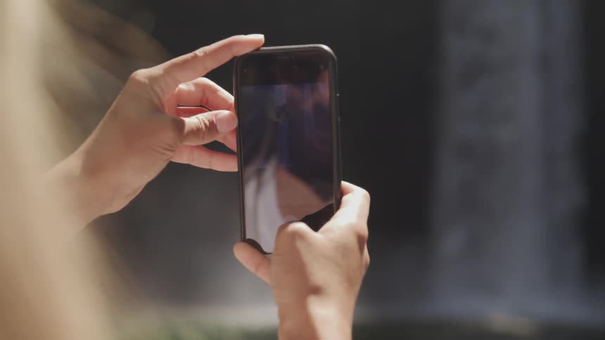 A woman holds a smart phone to take a picture of a waterfall