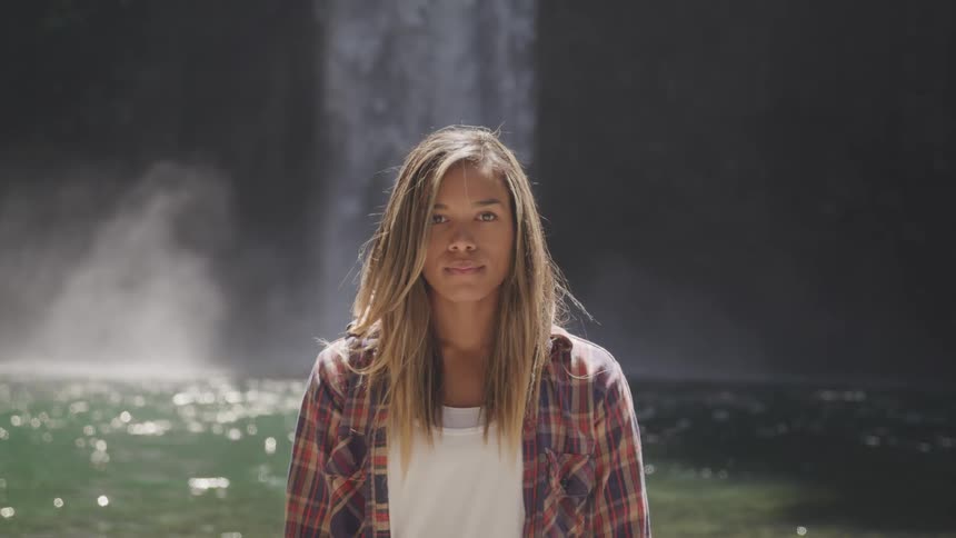 A girl stands in the middle of a lake beneath a waterfall