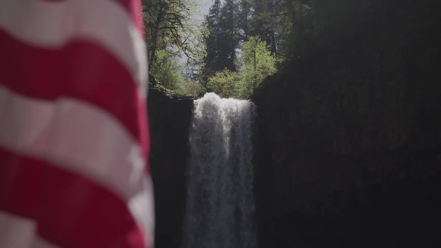 A person draped in an american flag stands on rocks next to a waterfall
