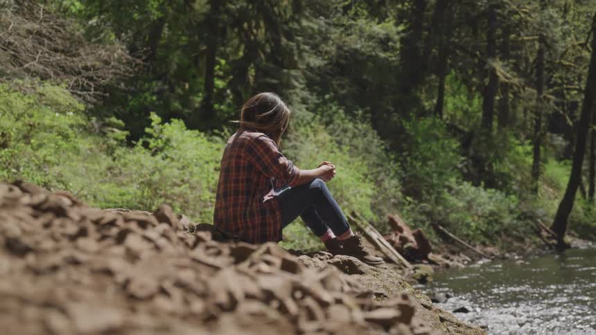 Woman wearing striped shirt sits on stones next to stream in the forest on a sunny day
