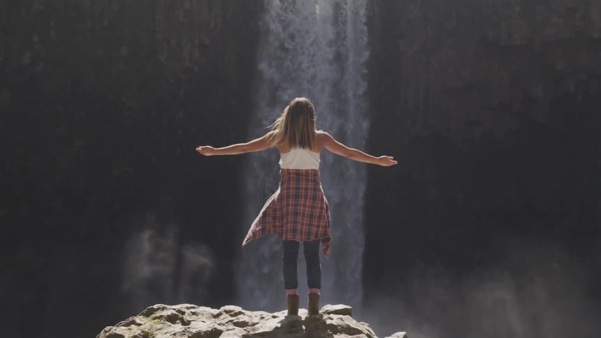 A woman standing on rock at the base of a waterfall raises her arms to the sky