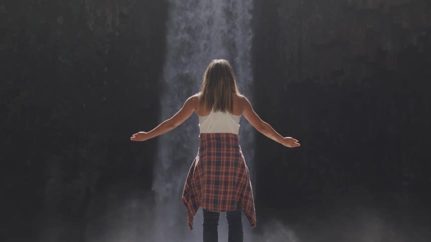A girl stands in front of a waterfall and raises her hands in the air
