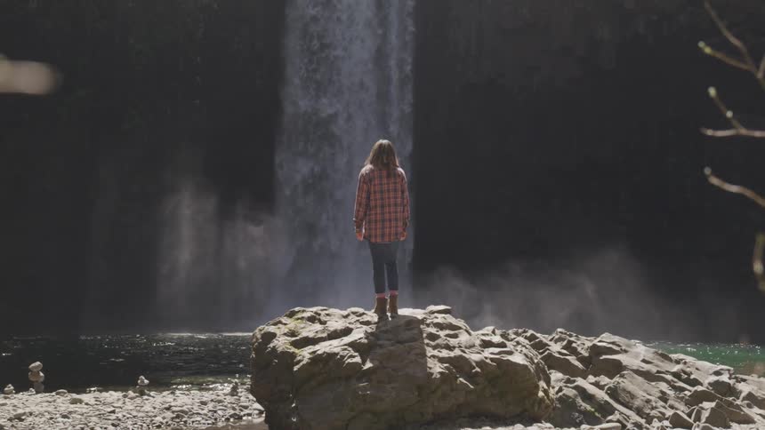 A white woman stands on a rock by a lake and watches a waterfall