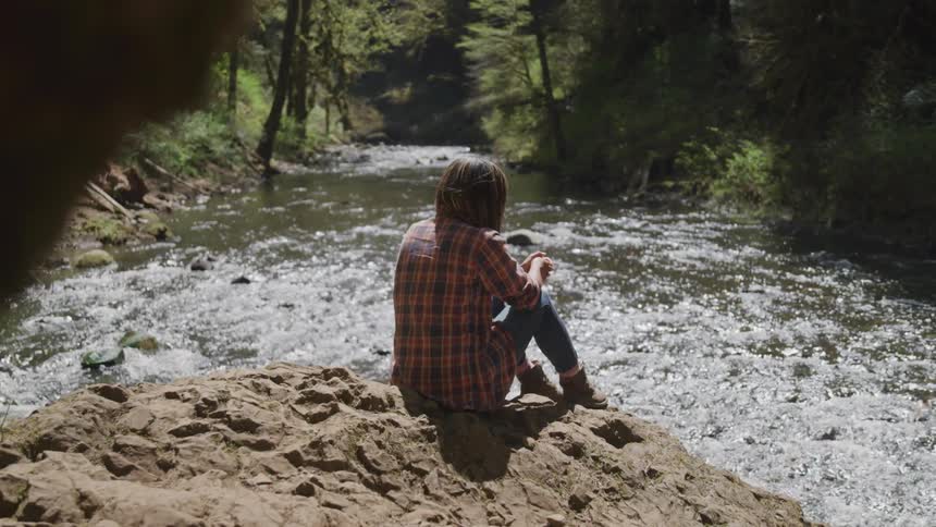 A young woman in a flannel shirt sits on a rocky ridge above a river flowing through a forest