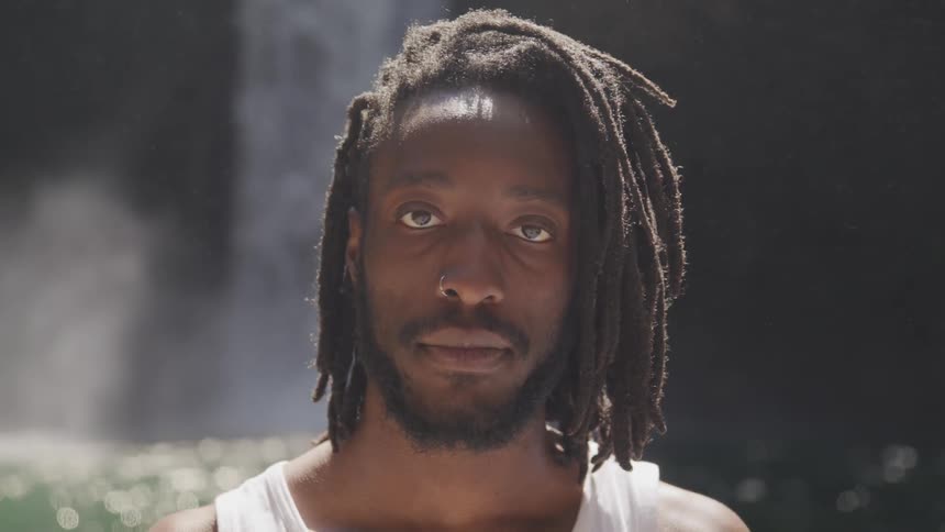 A black man sits in front of a waterfall and looks forward