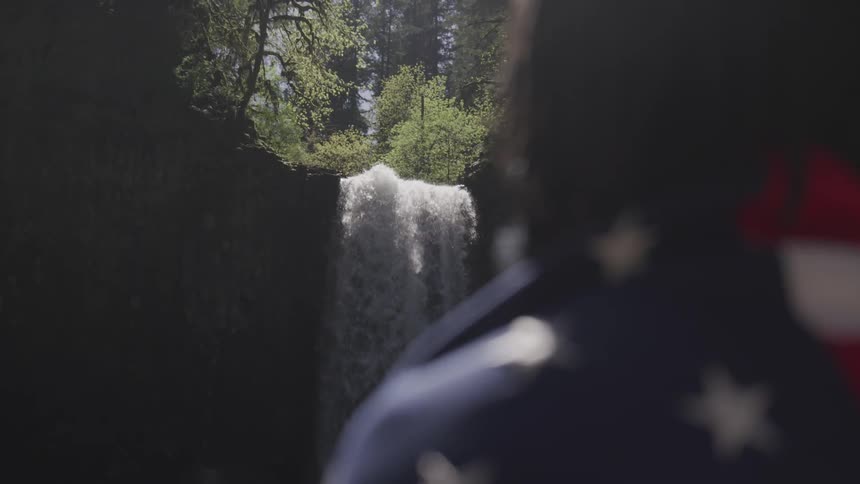 A person stands beside a waterfall with an american flag draped around their shoulders
