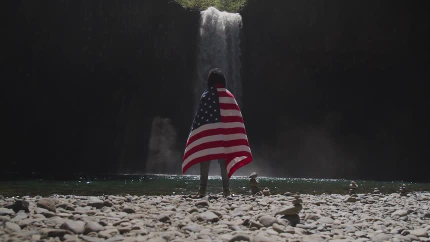 A woman draped in an american flag stands on rocks next to a waterfall