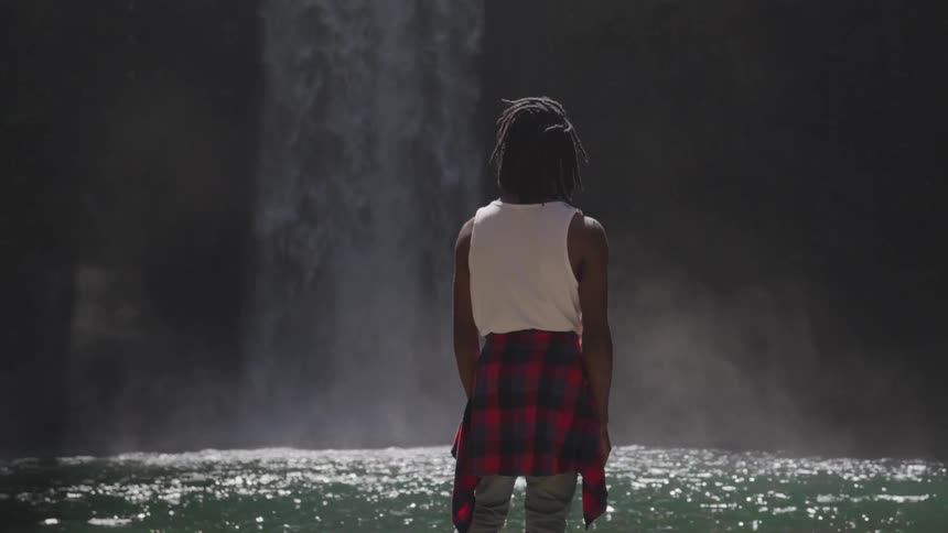 A person with a plaid shirt tied around their waist looks at a waterfall