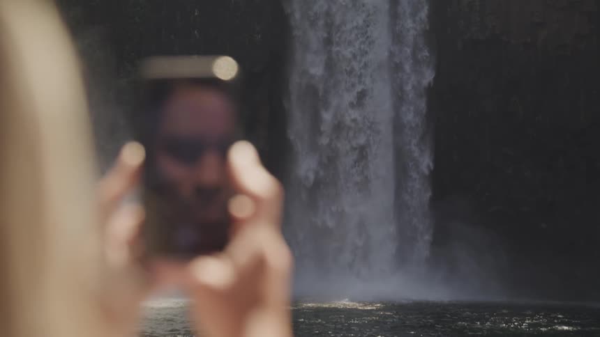 A woman is taking pictures of a waterfall with her phone