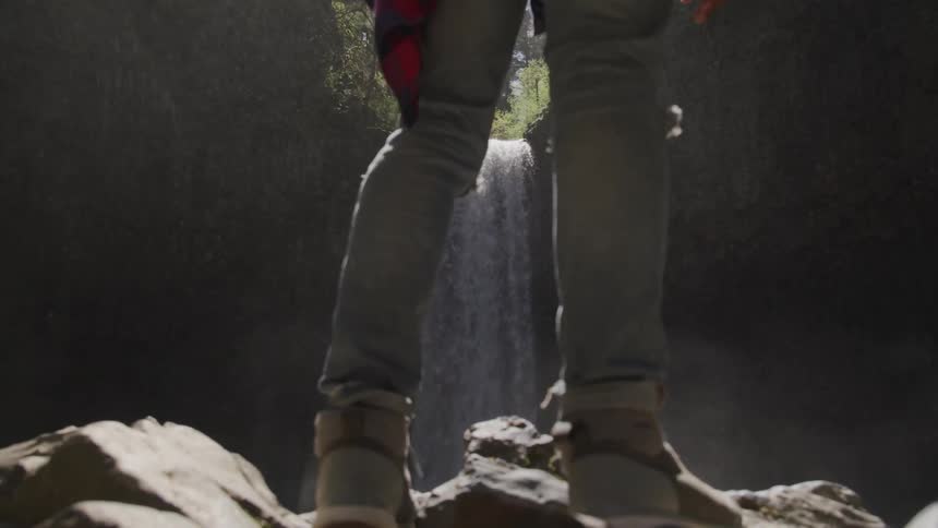 A hiker stands at the rocky base of a waterfall as water mists around them