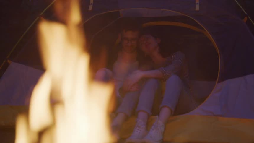 Man and woman sit in a tent next to the campfire in the forest at dusk