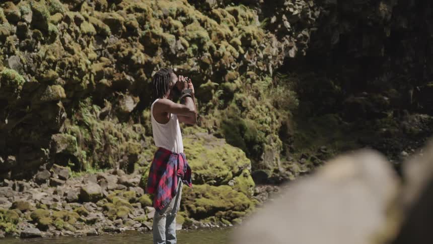Black man with dreadlocks takes a picture of waterfall next to the stream in the forest