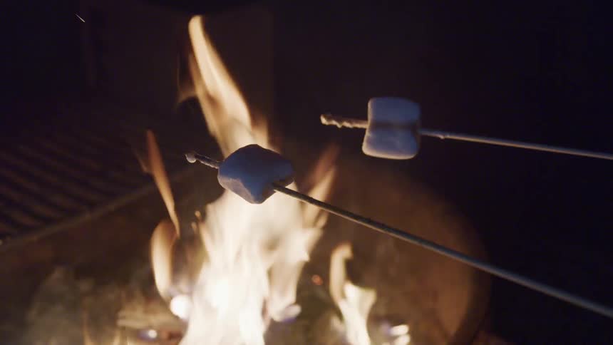 Two sticks with marshmallows on them are held over an open flame at night