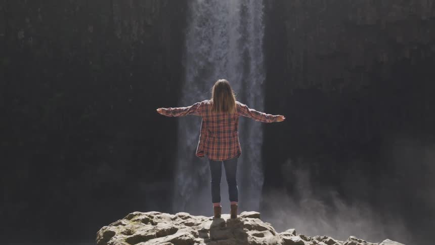 A woman in a plaid shirt stretches out her arms while standing in front of a waterfall