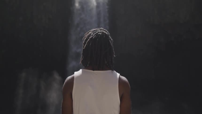 A young black man stands and looks at a water fall