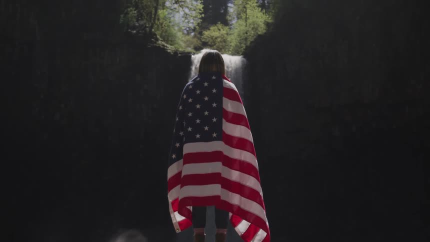 Person draped in american flag stands in cave looking at waterfall falling from outside